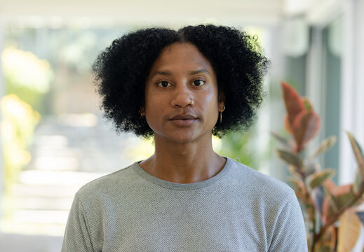 African American man posing by windows, gray longsleeve shirt, potted plants, nose hoop and hoops