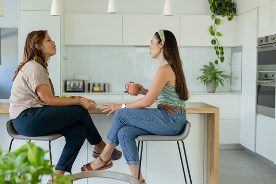 Two female friends sitting on stools at wooden kitchen island in modern kitchen holding ceramic mug