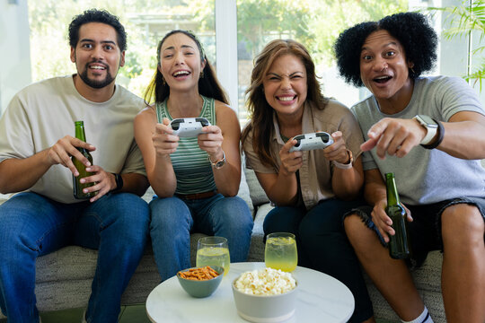 Diverse friends sitting on gray sofa playing games with white controllers holding bottles