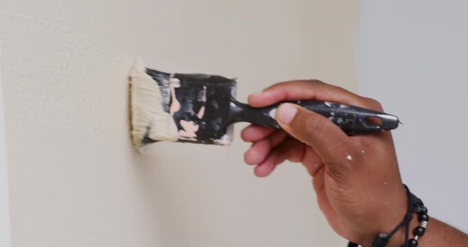 Male dipping brush into black tray, applying beige paint on drywall corner for even coverage