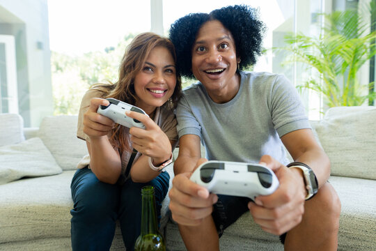 African American couple playing on sofa in living room holding white controllers near green bottle