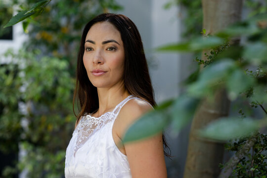 Adult female posing, looking left in garden wearing white lace dress amid foliage, tree trunk, wall