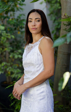 Woman standing beside tree trunk in garden wearing white lace embroidered dress and ring