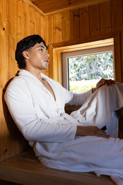 Asian man sitting on wood-paneled sauna bench by small window, wearing white bathrobe with belt