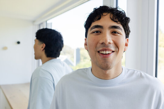 Adult Asian male standing by large windows reflecting in glass and wood counter, wearing light tee