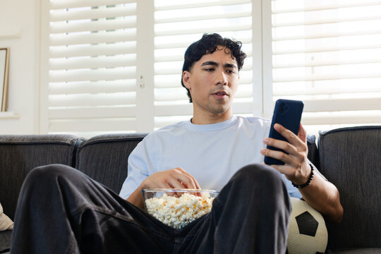 Asian man in blue tee watching smartphone while grabbing popcorn on sofa, soccer ball visible