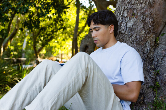 Asian adult man sitting in park leaning against tree trunk while using tablet wearing khaki pants