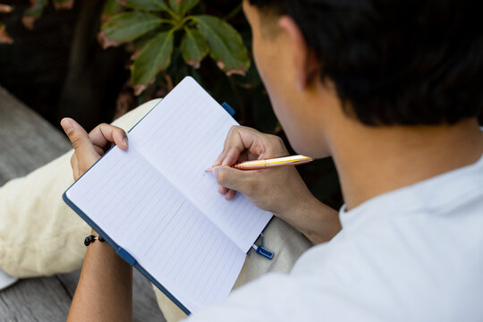 Asian adult male sitting on slatted bench in garden holding pen over lined notebook, copy space