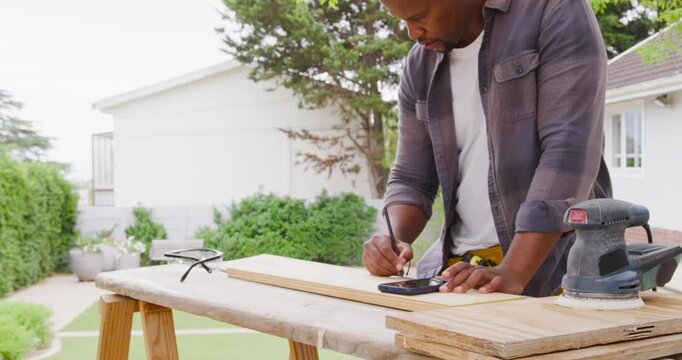 Placing plank on sawhorses African man wearing tool belt marking for cutting checking phone in yard