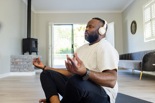 African mid adult man sitting cross legged on yoga mat in living room meditating with headphones
