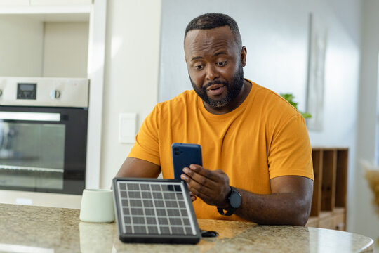 Mid adult African American man using blue smartphone beside portable solar panel on kitchen counter