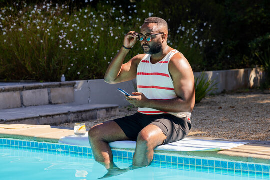 Mid-adult African American man sitting on pool coping with phone adjusting sunglasses near towel