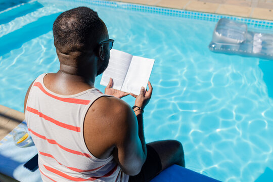 Man sitting poolside reading paperback on blue towel with smartphone, wearing striped tank top