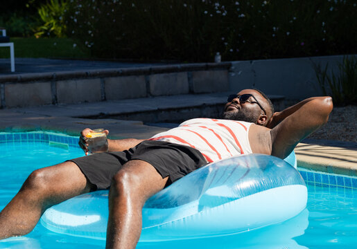 African American man reclining on float in backyard pool holding citrus drink wearing striped tank