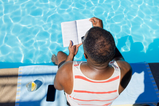 African American man sitting poolside reading book on towel, wearing striped tank and swim trunks