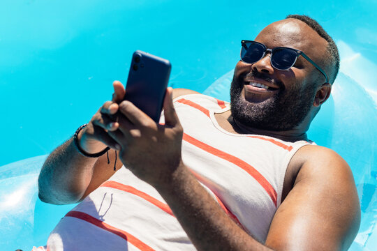 African American man lounging on float in blue water wearing shades striped tank holding phone