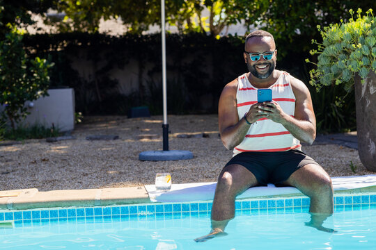 Mid-adult African man sitting on pool edge wearing striped tank top, sunglasses, holding smartphone