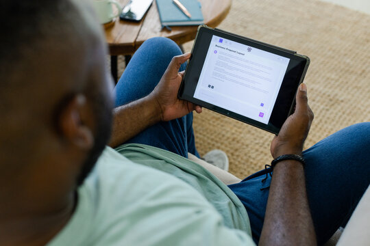 Mid-adult man sitting on sofa wearing light green T-shirt and blue jeans reading tablet proposal