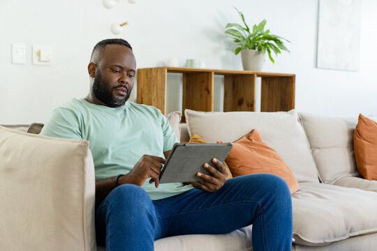 African American man sitting on sofa holding tablet in folio case, tapping screen in living room