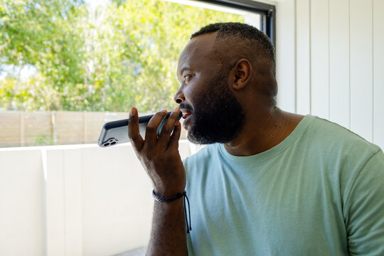 African American middle-aged man holding smartphone to mouth, facing open window, copy space