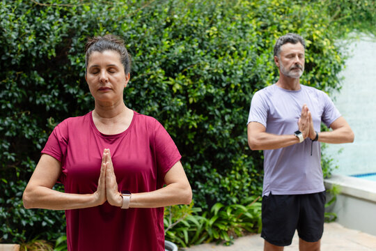 Couple practicing meditation standing on paved patio by raised pool edge wearing smartwatches