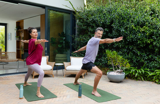 Diverse couple practicing standing yoga in gym clothes on patio with green mats and water bottles