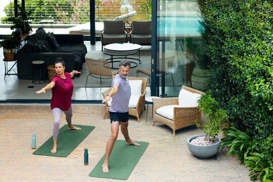 Senior couple doing yoga in activewear on green mats on brick patio near sliding glass doors