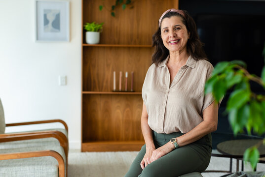Mature adult female sitting on upholstered stool in living room, smiling, hands resting on knee