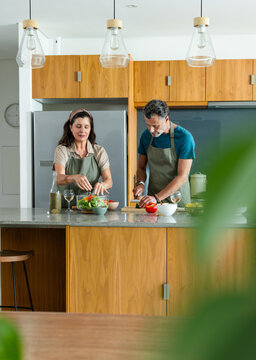 Senior couple wearing aprons, woman assembling salad on marble counter, man slicing pepper on board