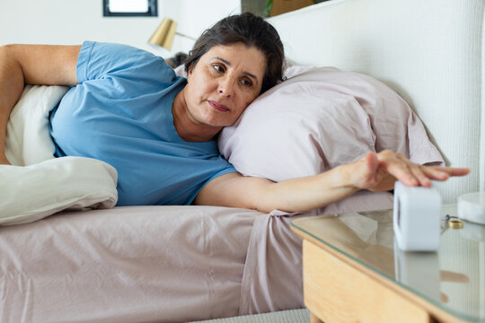 Mature woman reclining in bed wearing light blue shirt, reaching toward white digital alarm clock