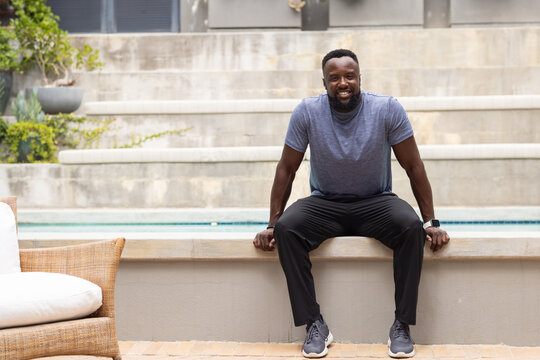 Male sitting on pool ledge at backyard patio, showing smartwatch and gray T-shirt, smiling