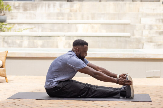 Adult African man stretching on black mat in patio by tiered seating holding sneakers wearing watch