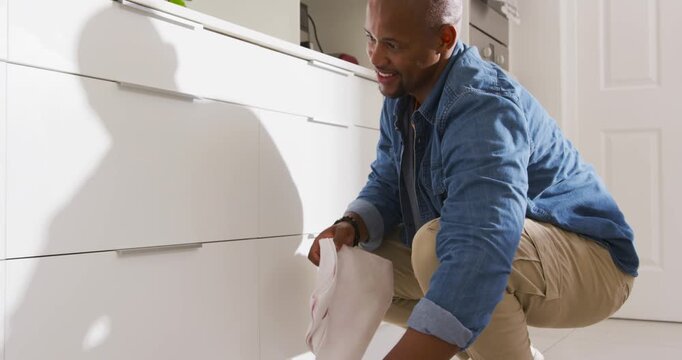 Man is unfolding white cloth, kneeling and wiping cabinet fronts in modern kitchen to remove marks