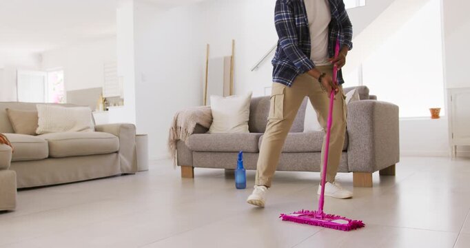 African American man in plaid spraying with blue bottle, pushing pink mop in living room smiling