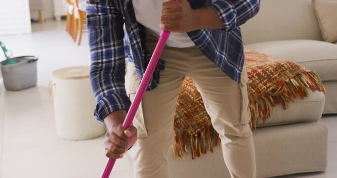 African American man in living room gripping pink mop, mopping tiled floor with blue spray bottle