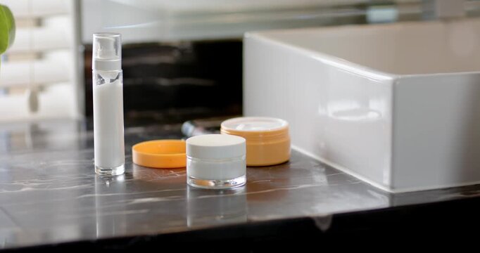 Skincare jars unmoved on marble sink, light shifting from blinds, sleeve entering then retracting