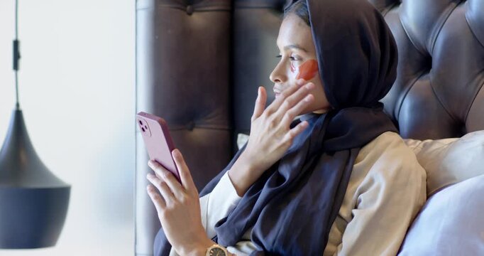 Placing red eye patch for eye care adult hijab-wearing female sitting in bedroom tapping pink phone