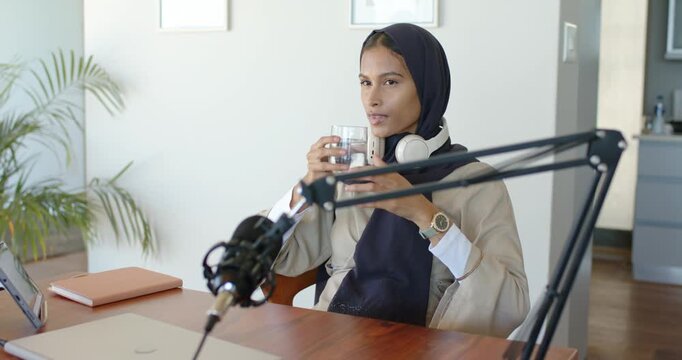 Woman pausing podcast at desk, reaching for clear water glass to hydrate, drinking, glancing at mic