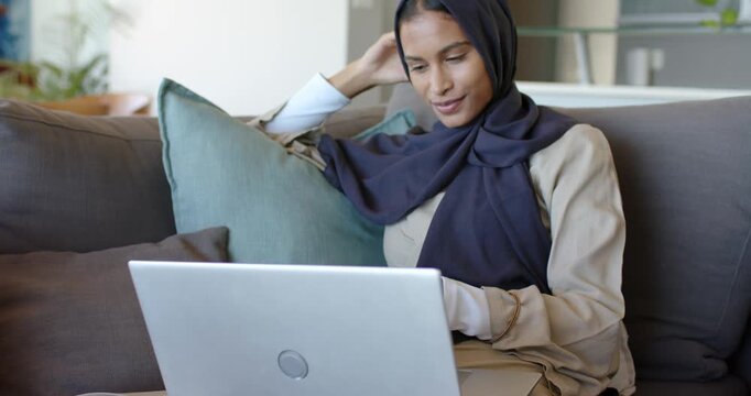 African American woman opening silver laptop, sitting on sofa at home, adjusting hijab and studying