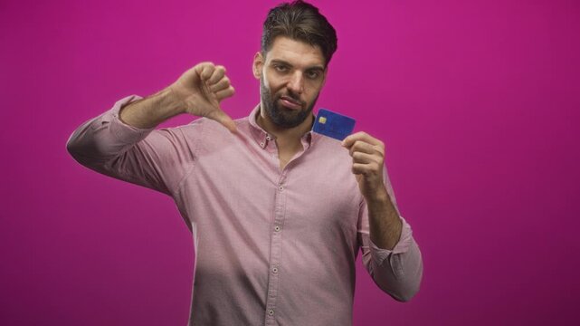 Hispanic man holding creditcard and showing thumbs down in studio with magenta backdrop; dissatisfaction trust concern.