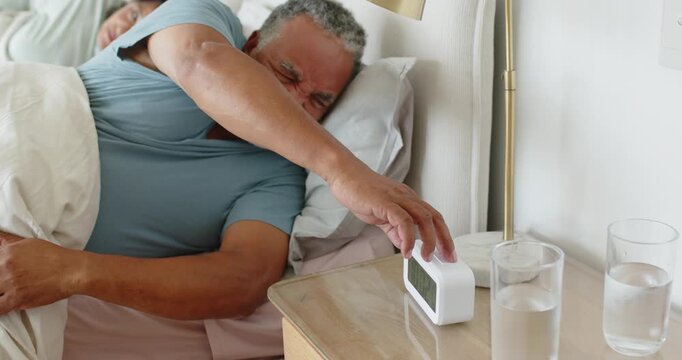 Alarm ringing Senior African American man in bed reaching clock silencing sitting as woman watching