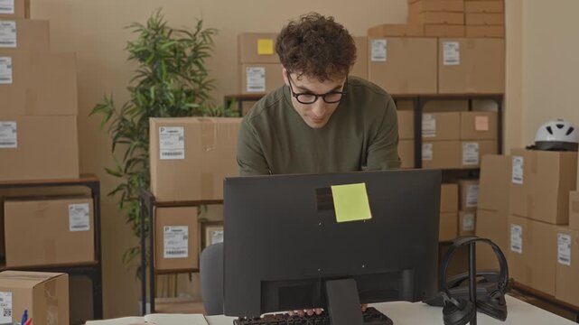Man types on computer amid stacked parcels, leaning over keyboard inside a packing building; small business focused.