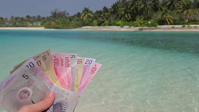 Stack of different denominations of Banknotes of Maldives, Maldivian rufiyaa in hand with maldivian island and ocean in background. Expensive travel concept