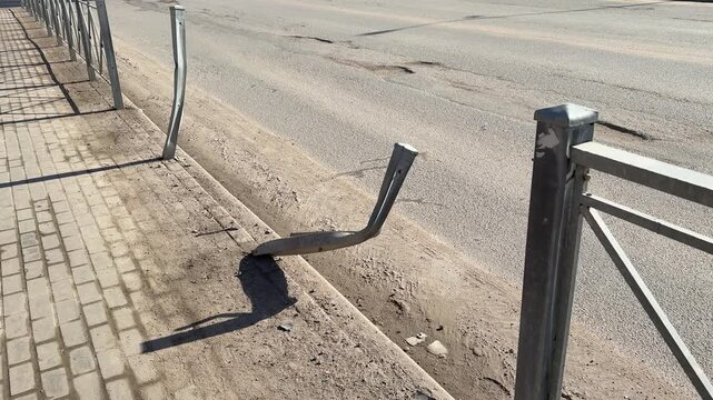 Bent fence post on sunny highway roadside twisted metal leaning over paved sidewalk, gravel scatter and scrape marks from impact.