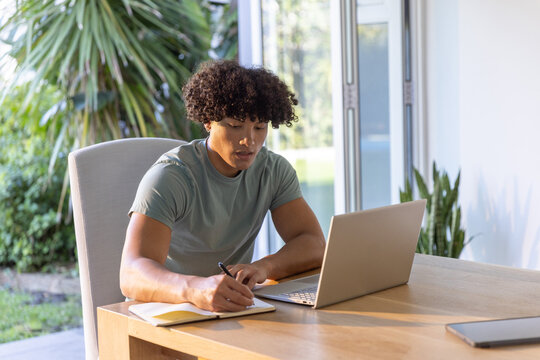 African American male writing in notebook with pen beside open laptop by sliding glass doors