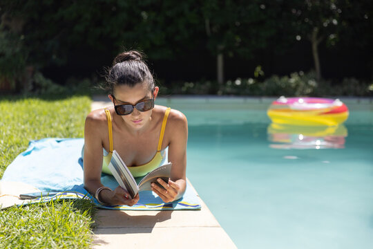Woman lying on stomach at pool reading book with shades, yellow straps, bright ring nearby