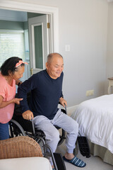 Senior Asian woman steadying and assisting Asian man transferring from wheelchair to bed in bedroom