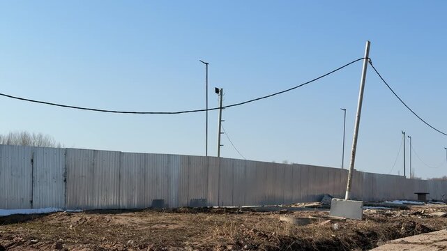 Construction site stainless steel fence against clear blue sky with lighting poles, utility wires, concrete bases and rough gravel.