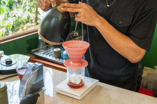 Barista hand pouring V60 pour over coffee with kettle into dripper over ice on scale, Japanese iced coffee flash brew method in natural light kitchen.