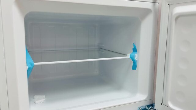 Empty freezer interior new refrigerator showing wire shelf with protective blue tape, open door, pristine white plastic walls.
